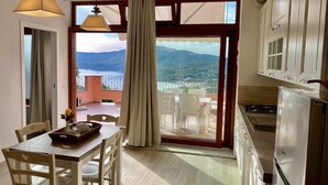 A dining area with a table and chairs, a view of the sea and mountains through large windows, and a kitchen with cabinets and a refrigerator. - Villetta Zuccale (Capoliveri)