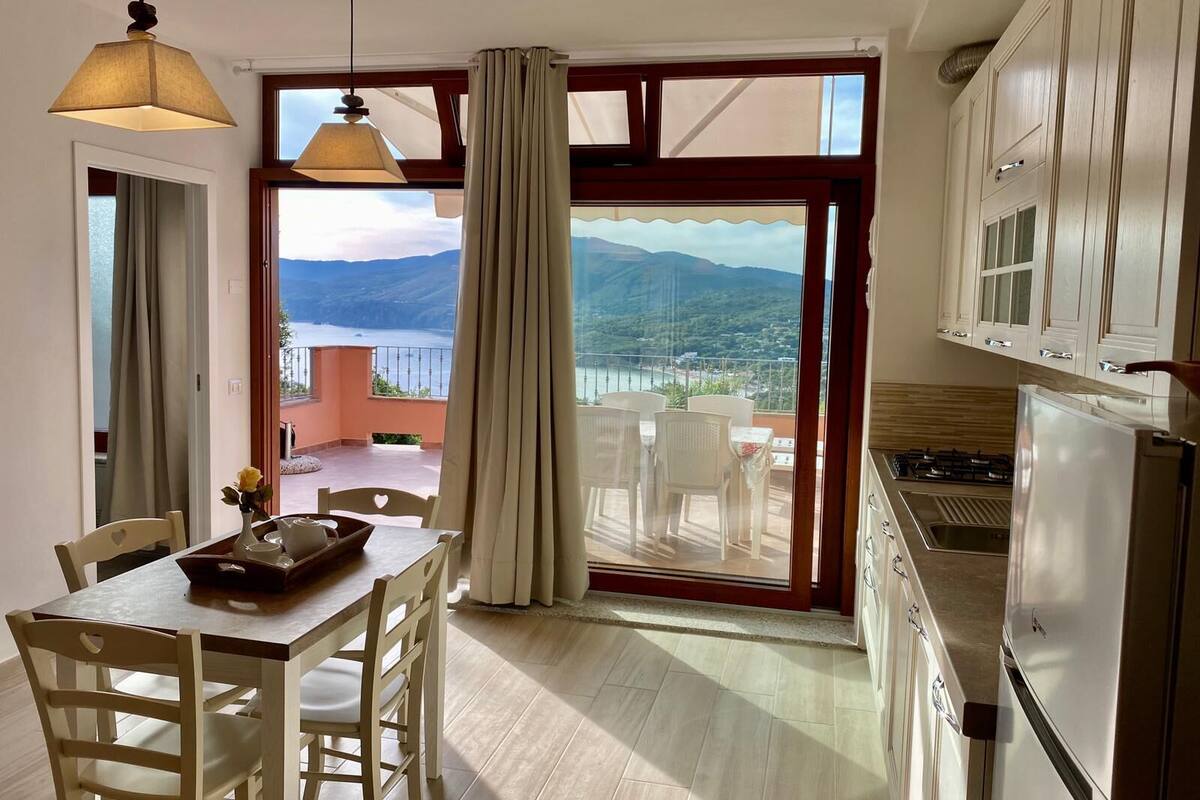 A dining area with a table and chairs, a view of the sea and mountains through large windows, and a kitchen with cabinets and a refrigerator.