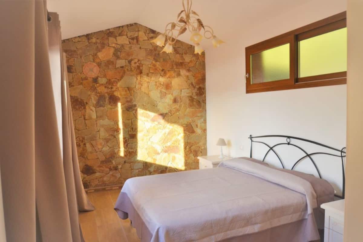 A bedroom with a stone wall, a bed with a metal frame, a window with blinds, and a nightstand with a lamp.