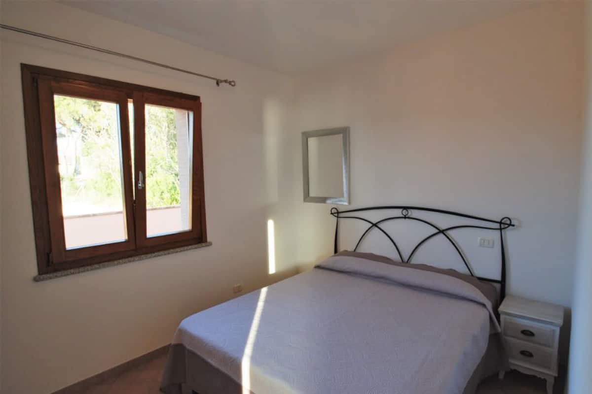 A bedroom with a bed, a nightstand, a window, and a mirror on the wall.