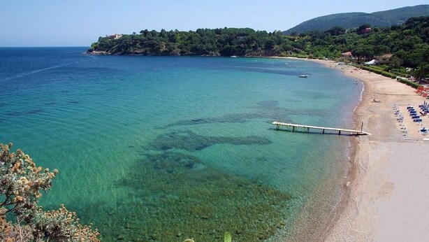 A coastal beach with clear waters, a pier, and a sandy shore with beach umbrellas. - VerdeBlu (Capoliveri)