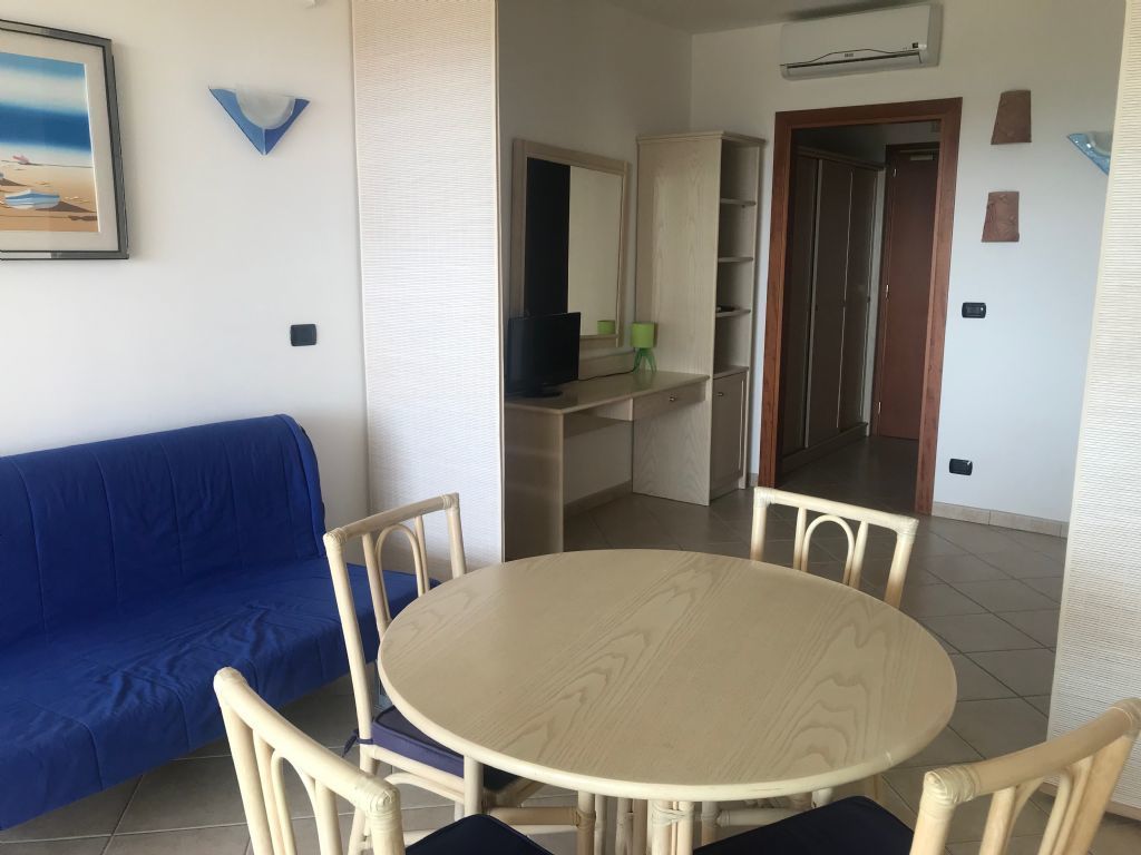 Una sala de estar con un sofá azul, una mesa redonda de madera y dos sillas. Hay un televisor sobre un mueble bajo, un espejo y un aparador con estantes.