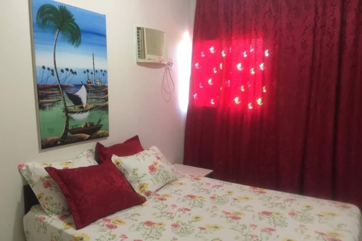 Une chambre à coucher avec un lit, des rideaux rouges, un climatiseur fixé au mur et un tableau représentant un paysage tropical.