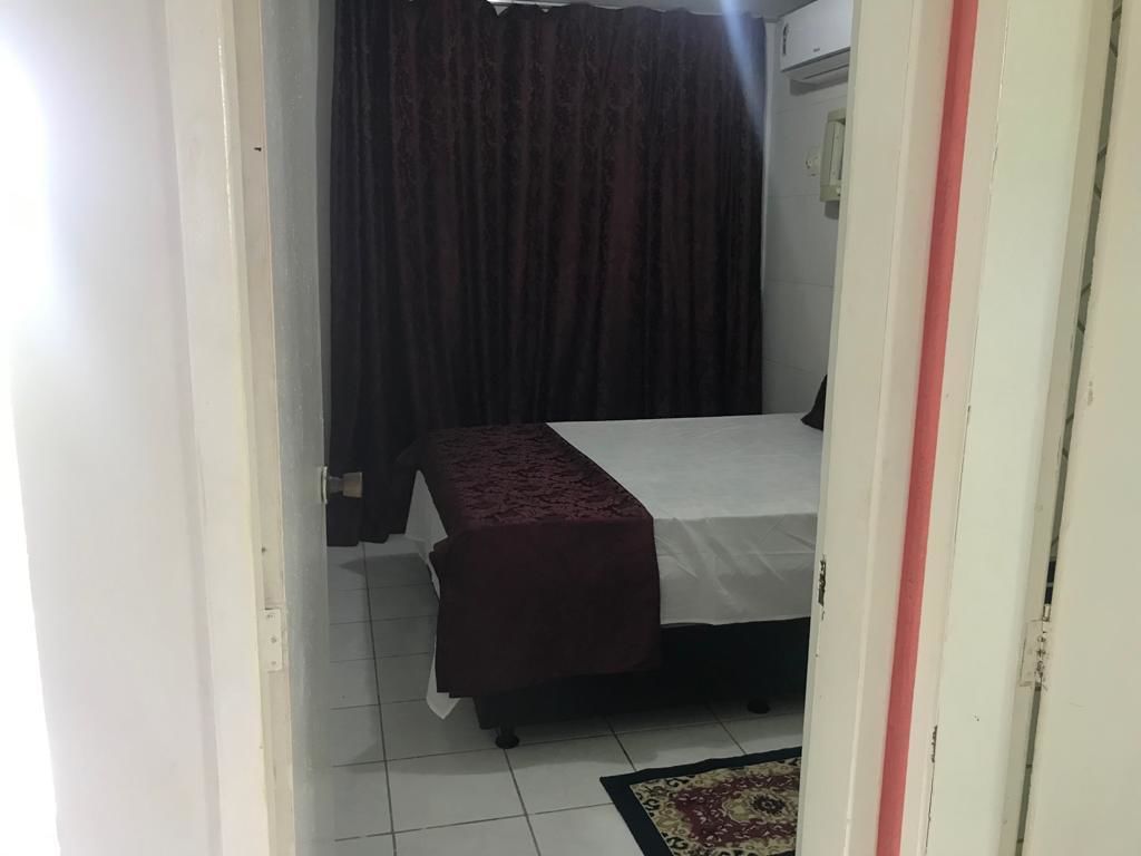 Una stanza piccola e scarsamente illuminata, con un letto singolo, una tenda scura e un pavimento piastrellato.