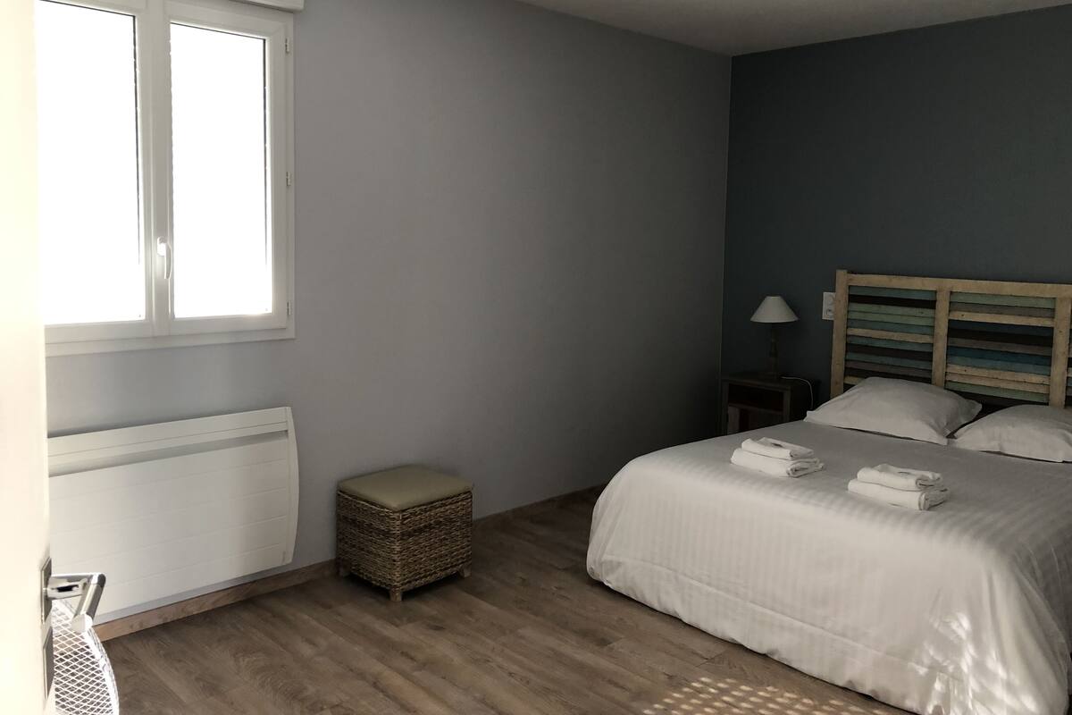 Ein Schlafzimmer mit einem Bett, einem Nachttisch, einer Lampe, einem Korbstuhl und einem Fenster.