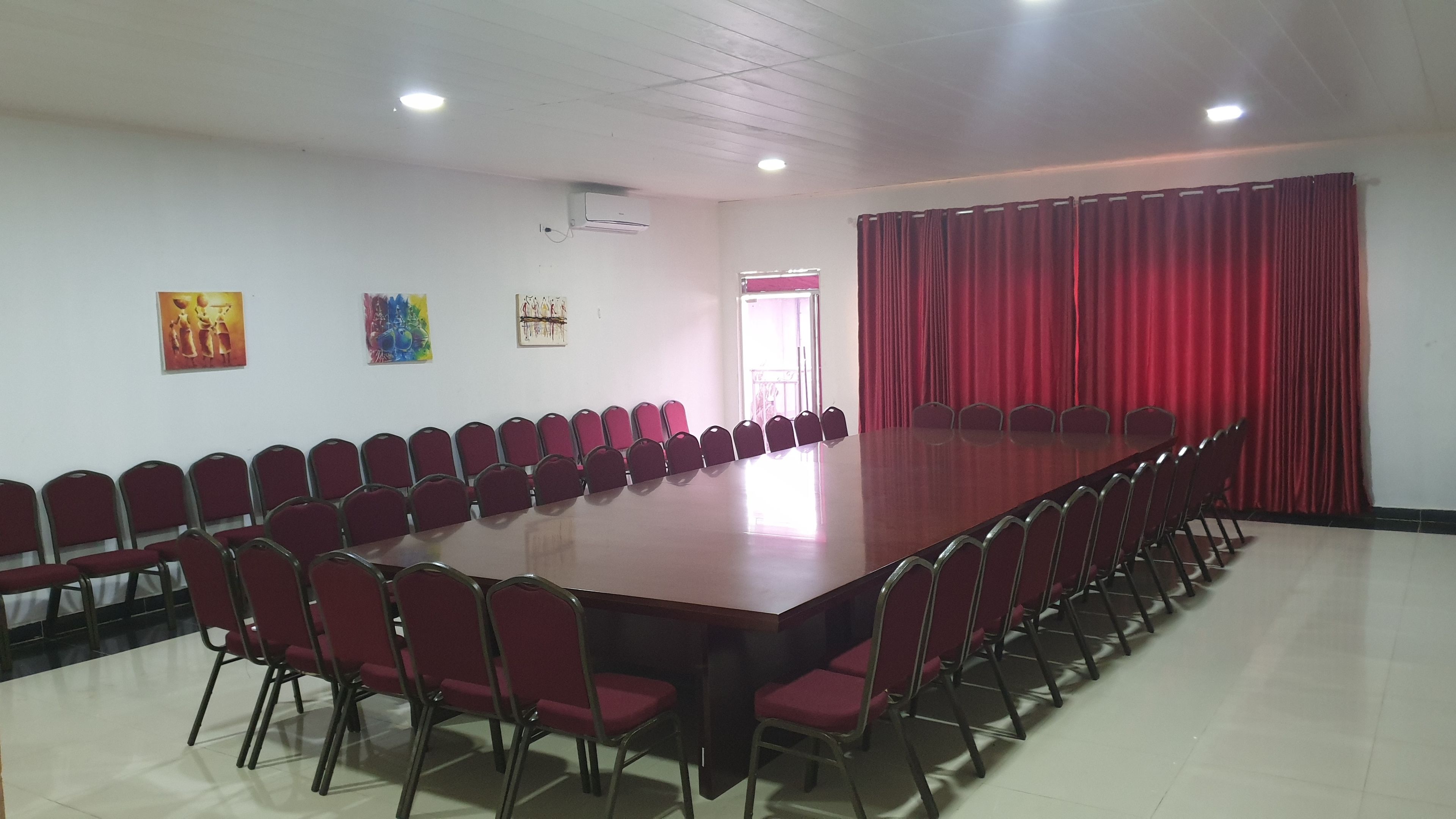 Un salón de conferencias con una larga mesa y filas de sillas, cortinas rojas y cuadros en la pared.