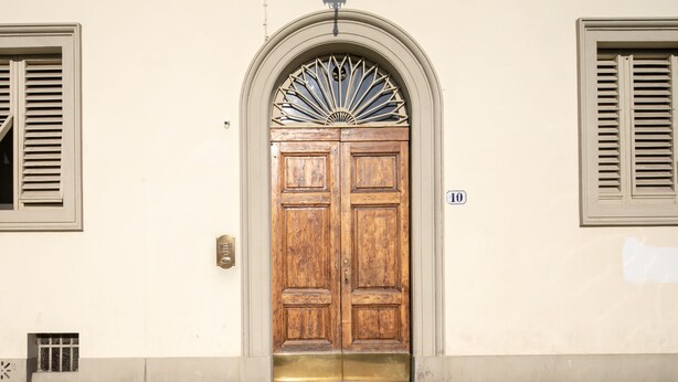 Eine Holztür mit dekorativem Bogen darüber, eingebaut in ein Gebäude mit beige gestrichenen Wänden und Fensterläden.