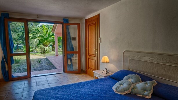 A bedroom with a bed, a nightstand, a lamp, and a sliding glass door leading to a garden. - Luisa (Capoliveri)