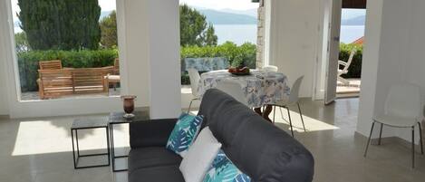 Una sala de estar moderna con un sofá, una mesa de centro y un comedor con una mesa y sillas. Se observa una vista de agua y montañas al fondo.