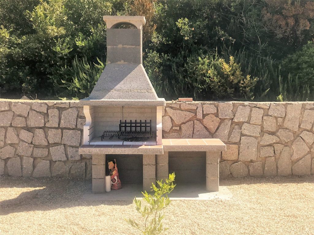 A stone barbecue grill with a chimney, set against a stone wall and surrounded by greenery.
