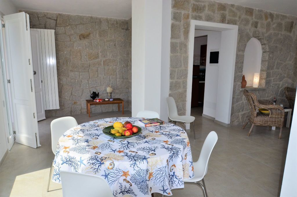 A dining area with a round table covered in a tablecloth, surrounded by white chairs. A fruit bowl is on the table, and there is a small table with a lamp and a book in the background.