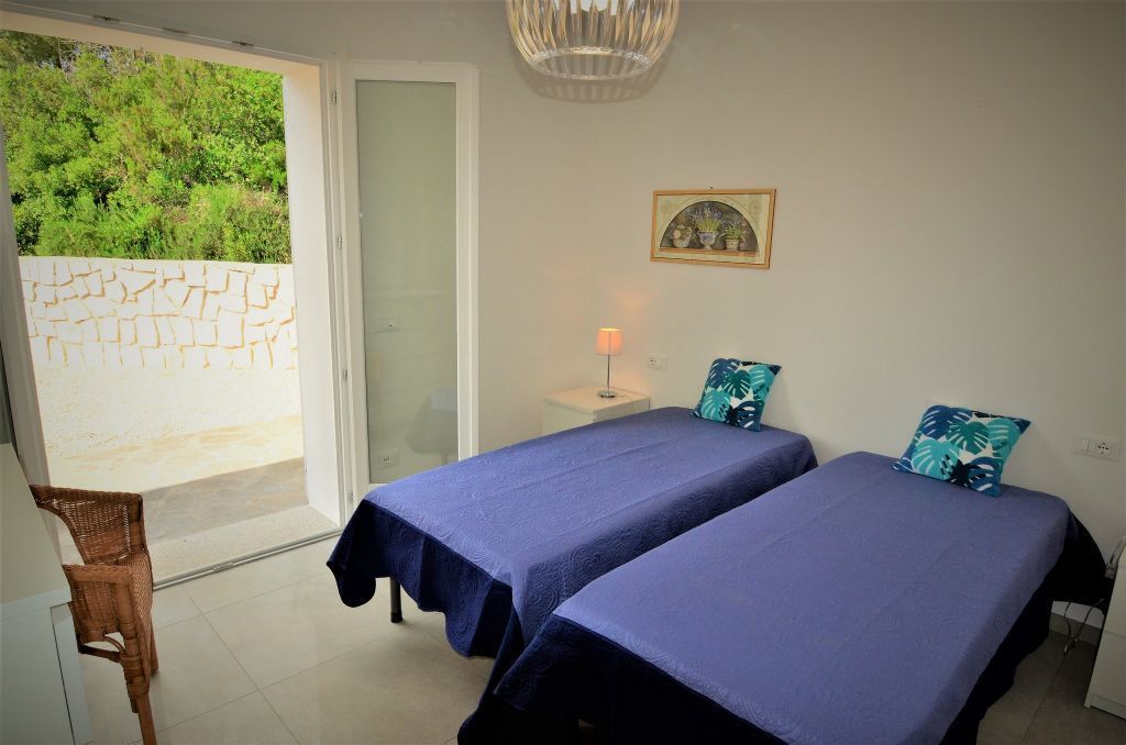 A room with two beds, a small table with a lamp, a wicker chair, and a view of greenery through a glass door.