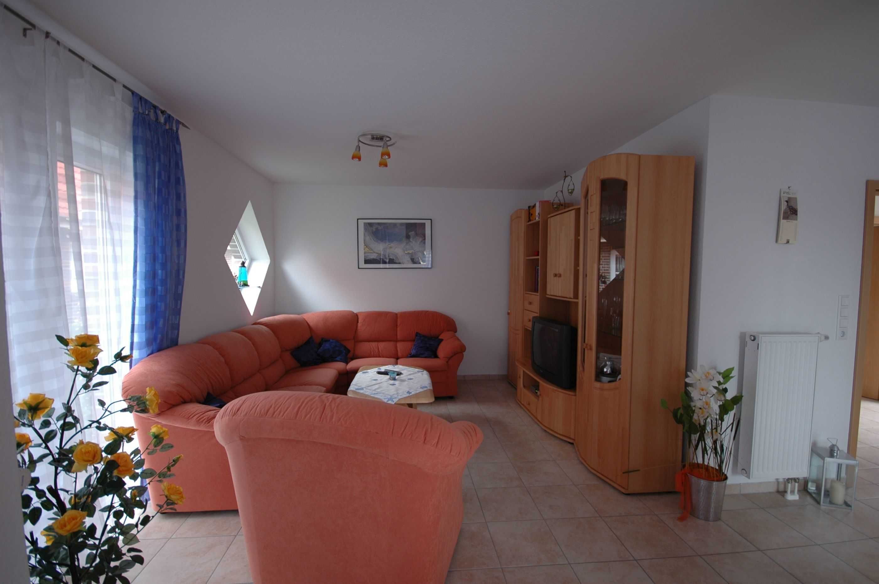 A living room with a large orange sofa, a small table, a wooden cabinet with a television, and a window with blue curtains.