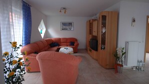Una sala de estar con un sofá grande color naranja, una mesita, un mueble de madera con un televisor y una ventana con cortinas azules.