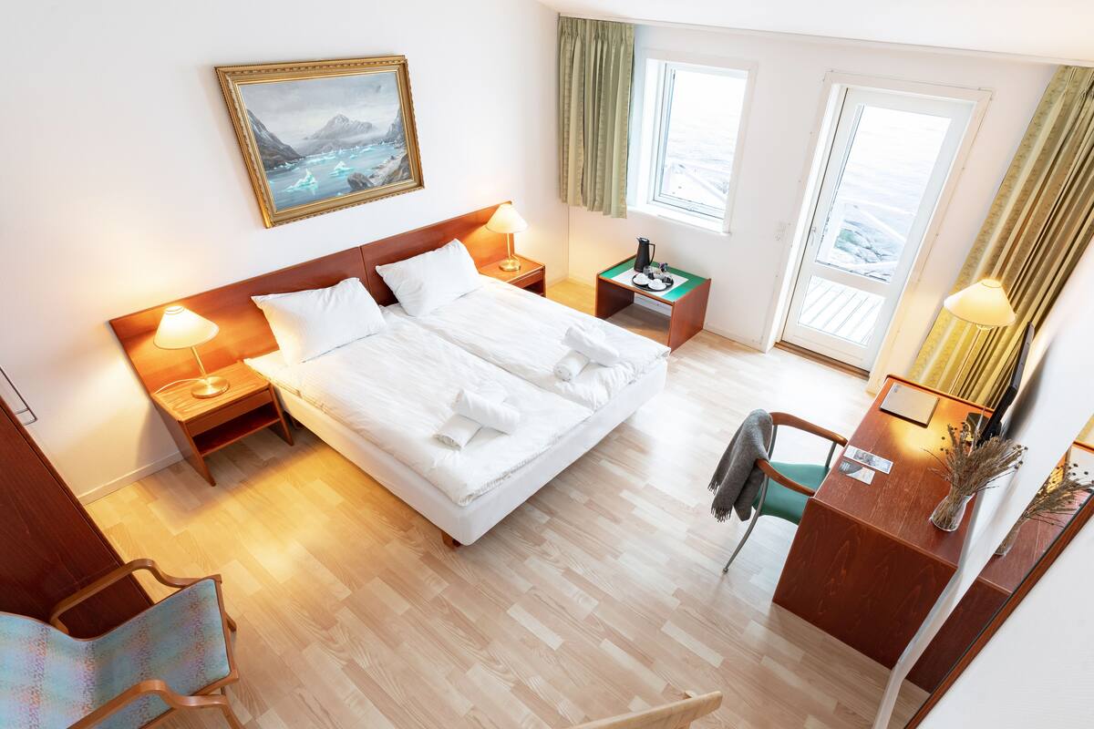 Een hotelkamer met een groot bed, twee nachtkastjes, een bureau met stoel, een spiegel en een raam met gordijnen.