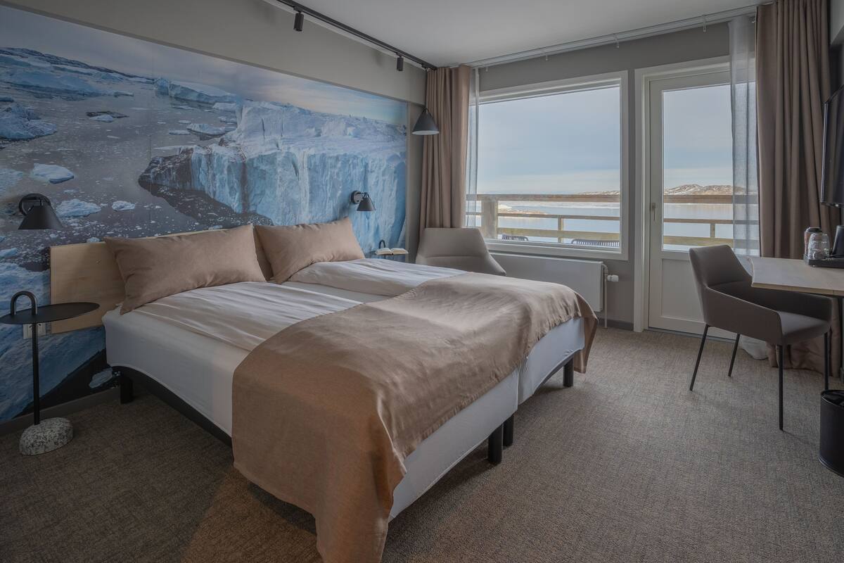 Een hotelkamer met een bed, een bureau, een stoel, een groot raam met uitzicht op zee, en een muurschildering van een gletsjer.