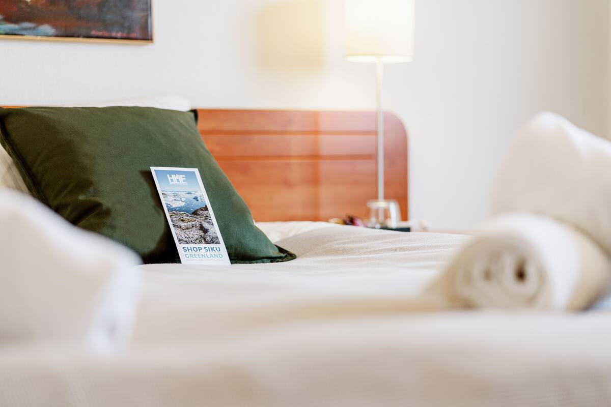 Een net opgemaakt bed met een groen kussen en een opgerolde handdoek, een brochure op het kussen, en een lamp op de achtergrond.