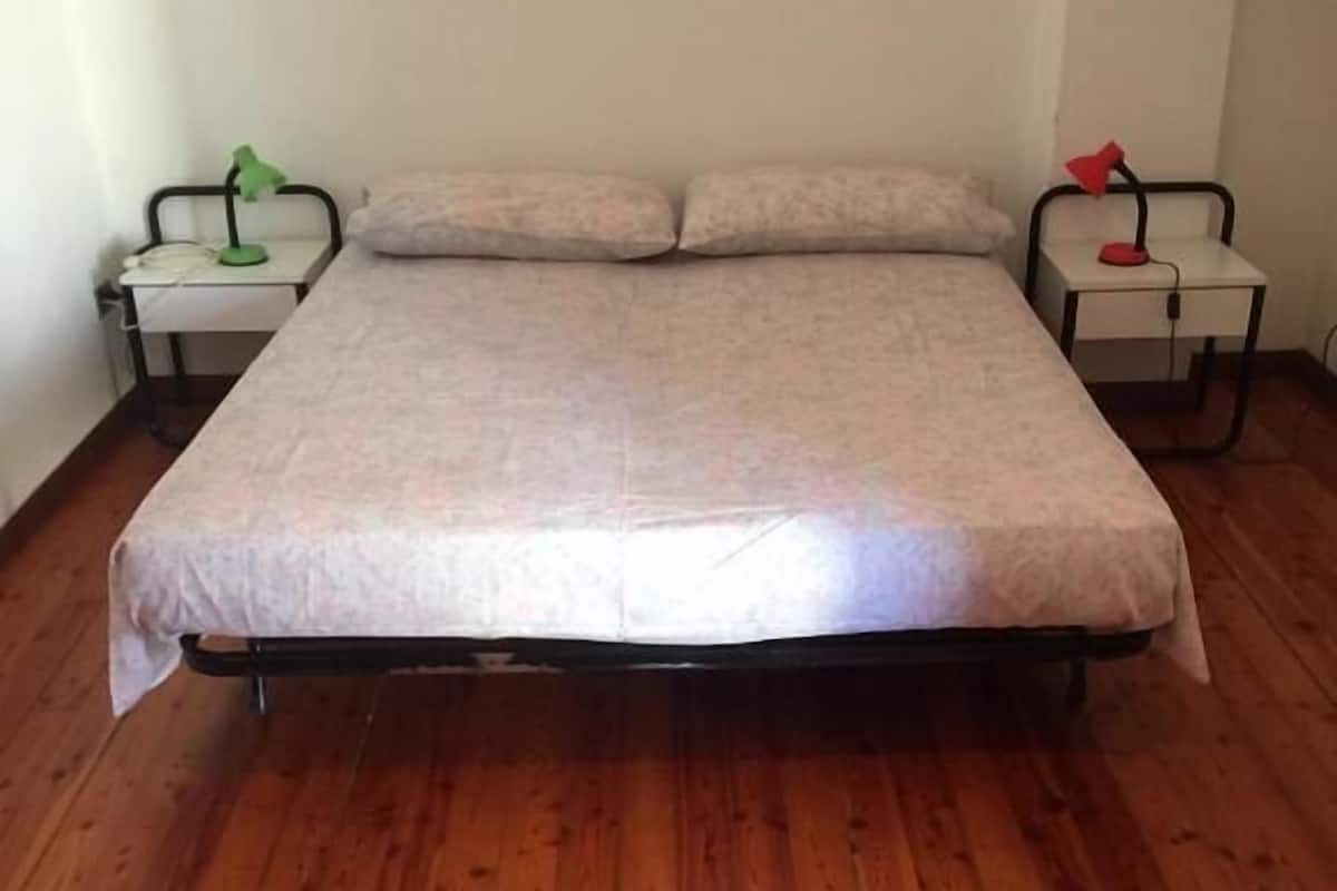 A single-bed room with a wooden floor, a bed with a white mattress and matching pillows, two bedside tables with lamps, and a white wall.