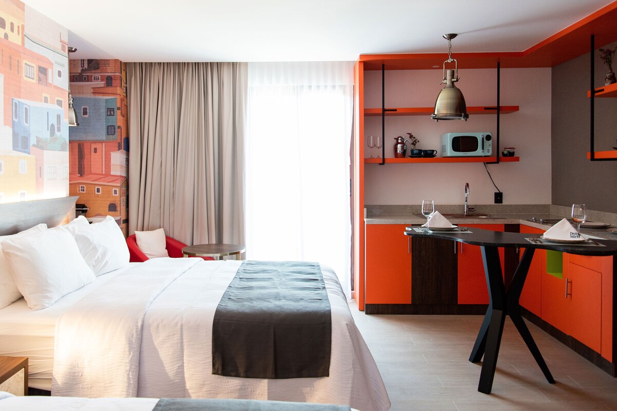 A hotel room with a bed, a desk, a kitchenette, and a view of buildings through the window.