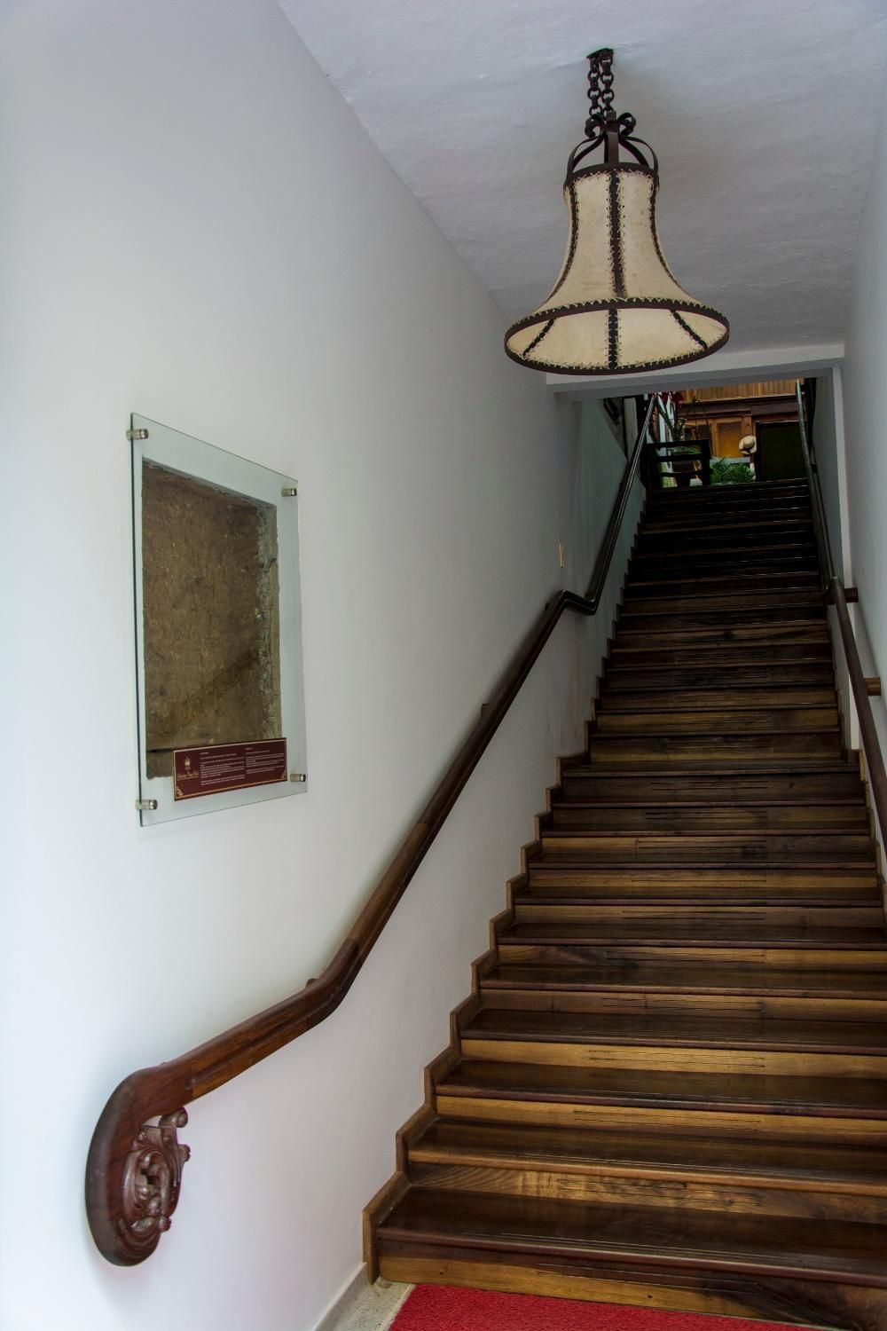 Una escalera de madera con barandilla, una lámpara colgante y un aviso en el pared.