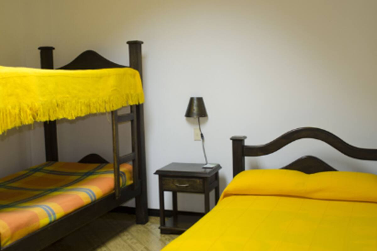 A room with two beds, one with a yellow bedspread and the other with a patterned bedspread. There is a small table with a lamp between the beds.