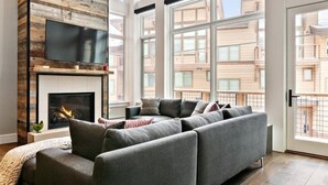 Ein modernes Wohnzimmer mit Ecksofa, Kamin, Flachbildfernseher und großen Fenstern mit Blick auf die Stadt.
