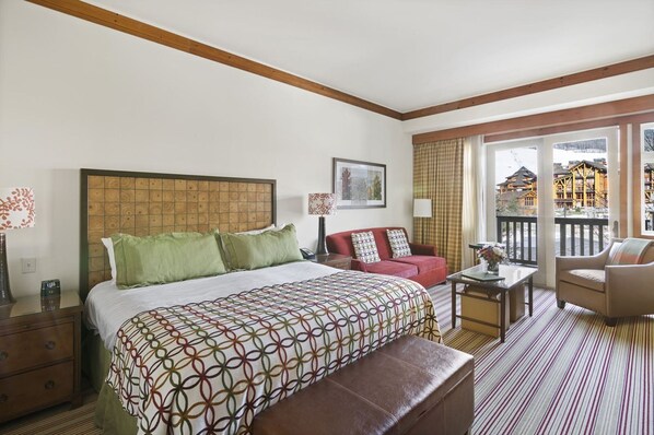 A hotel room with a large bed, a red sofa, a small table, and a balcony with a view of buildings. - 1228/1230 Double Studio @ The Lodge at Spruce Peak (Stowe)