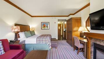 A hotel room with a bed, a desk, a TV, a fireplace, and a hallway leading to the bathroom.