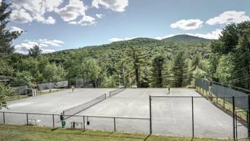 An outdoor tennis court surrounded by a fence, with a mountainous backdrop and trees.