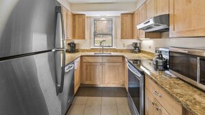 A compact kitchen with wooden cabinets, stainless steel appliances, and a window with blinds. - Lodge Condo 30 (Stowe)