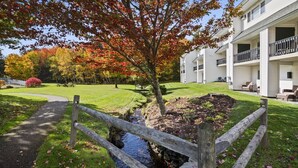A residential building with a small stream, a wooden bridge, and a lawn with autumn foliage. - Lodge Condo 30 (Stowe)