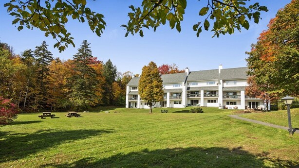 A large, white building with multiple balconies and a well-maintained lawn with picnic tables. - Lodge Condo 30 (Stowe)