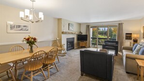 A spacious living room with a fireplace, a dining area with wooden chairs, and a large window offering a view of the outdoors. - Lodge Condo 30 (Stowe)