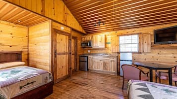 A cabin with wooden walls and floors, a kitchen with a microwave and refrigerator, a dining area with a table and chairs, and a bed with a quilt.