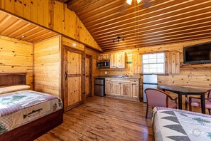 A cabin with wooden walls and floors, a kitchen with a microwave and refrigerator, a dining area with a table and chairs, and a bed with a quilt.