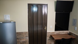 A room with a wooden door, a small refrigerator, a brick accent wall, a wooden table, and a black chair. - Hotel Nixon (Itai)