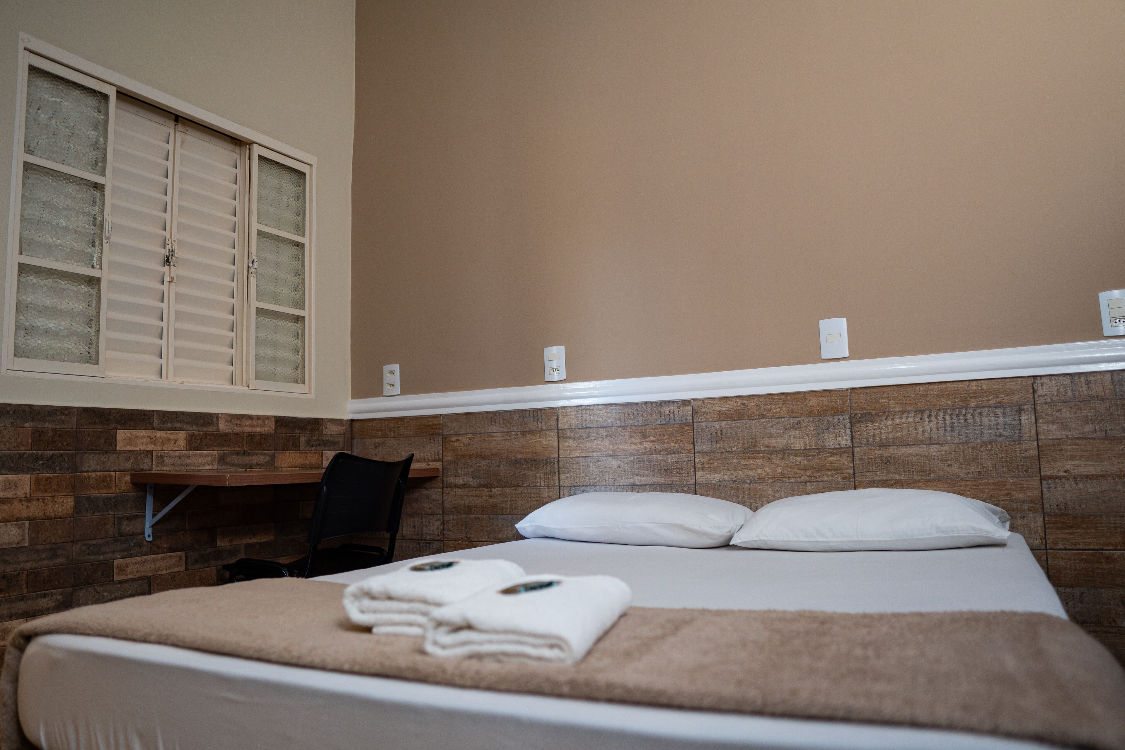 A hotel room with a bed, a wooden headboard, a window with blinds, a chair, and a shelf with a light switch.