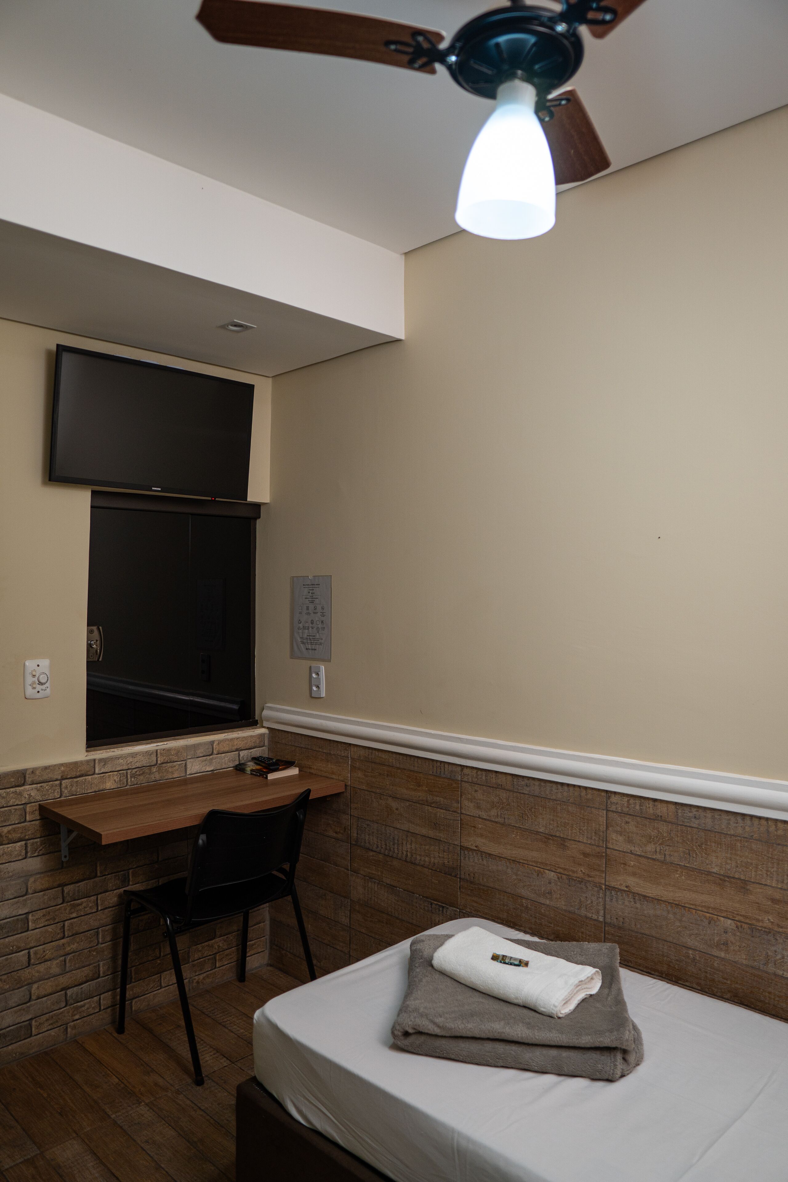 A hotel room with a bed, a desk, a chair, a TV mounted on the wall, and a ceiling fan.