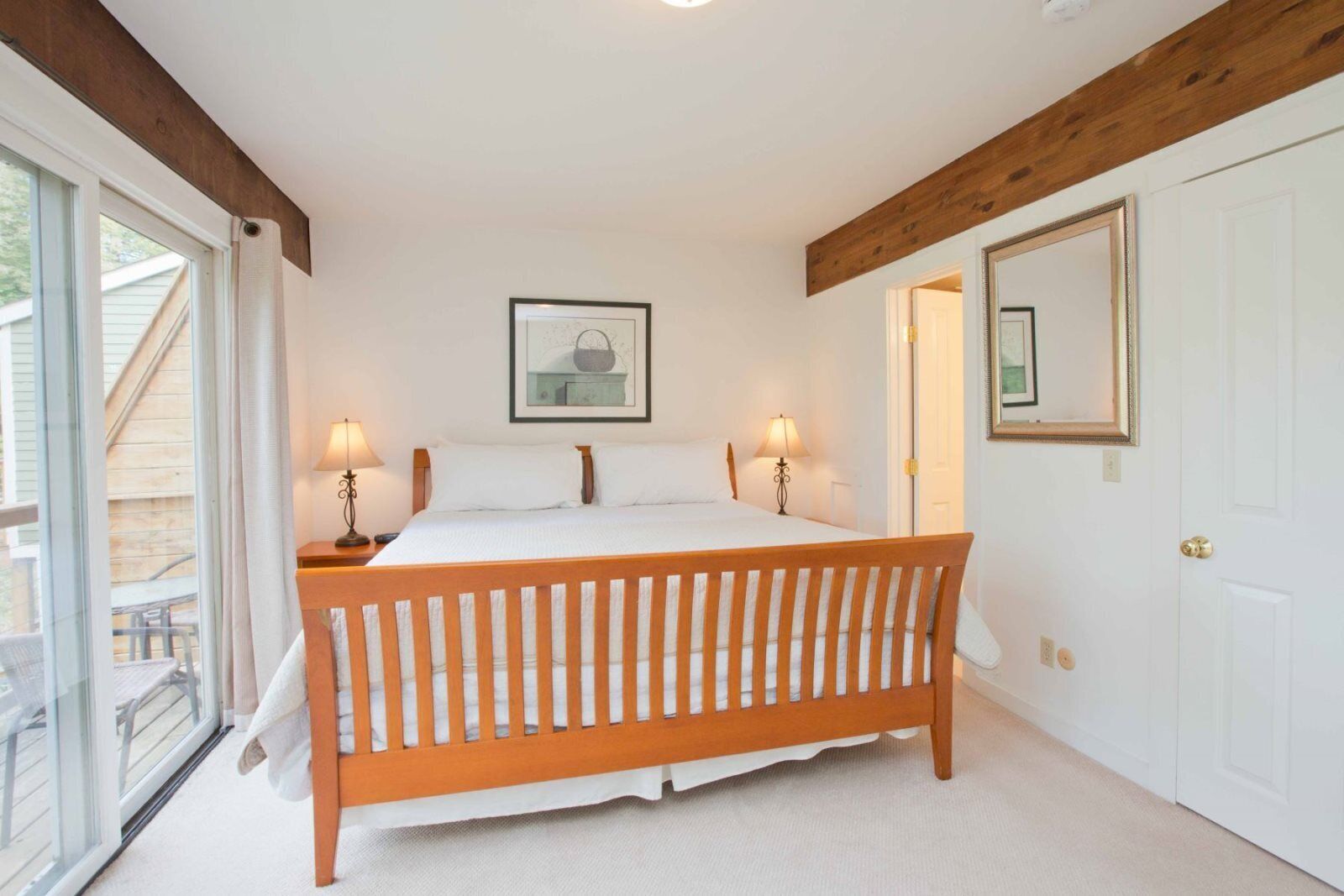A bedroom with a wooden bed, two bedside lamps, a framed picture on the wall, and a sliding glass door to the outside.