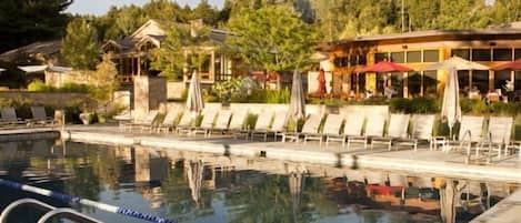 A pool area with lounge chairs and umbrellas, a building with outdoor seating, and a reflection in the water.