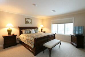 Une chambre à coucher comprenant un lit, deux tables de chevet avec des lampes, un banc, une commode, un téléviseur et une fenêtre avec des stores.