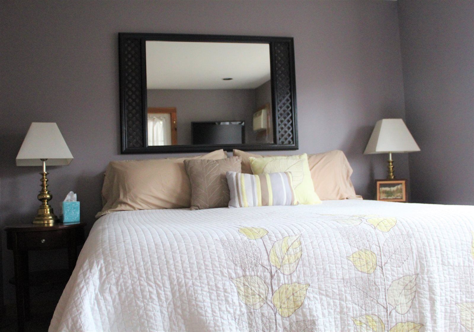 A neatly made bed with a leaf-patterned quilt, two bedside lamps, and a large mirror above the bed.