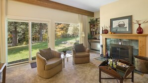 A cozy living room with a fireplace, a bookshelf, and a view of a garden through large sliding glass doors. - Brookside 2 BR at Topnotch (Stowe)