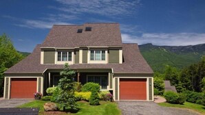A two-story house with a steeply pitched roof, two garage doors, and a well-maintained front yard with shrubs and a small tree. - 627a_topnotch (Stowe)