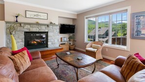 A living room with a stone fireplace, leather sofa, wicker chair, and a large window with a view of houses. - 608b_topnotch (Stowe)