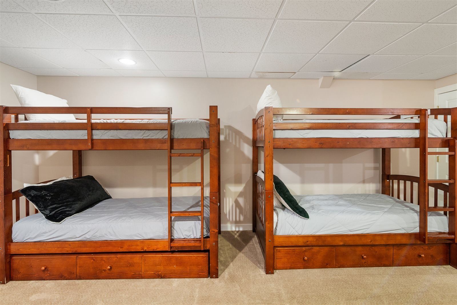 Two bunk beds with wooden frames and grey bedding, in a room with a tiled ceiling and carpeted floor.