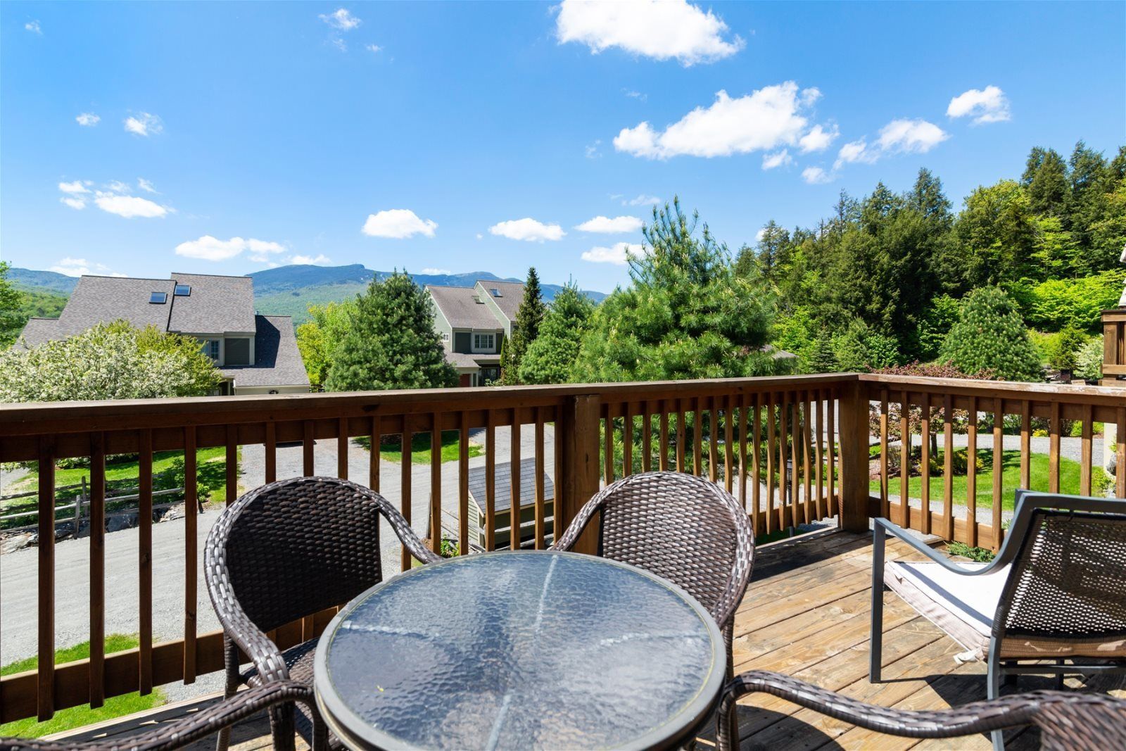Una terraza con sillas de mimbre y una mesa de vidrio, con vistas a una zona residencial con casas y árboles.