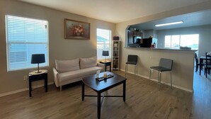 Ein Wohnzimmer mit Sofa, Couchtisch und einem Gemälde an der Wand. Es gibt eine Küchenzeile mit Mikrowelle und Spüle.
