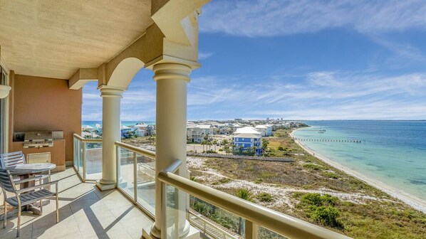 A balcony with a view of a beach and ocean, featuring a table and chairs, and a distant view of a resort area.