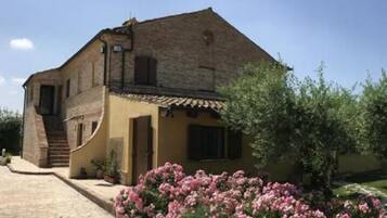 A rustic stone building with a sloped roof, a small staircase leading to the entrance, and a garden with pink flowers.